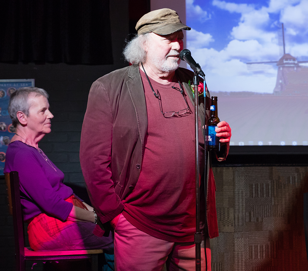 Larry Carius, dressed in maroon shirt with his wife Kay sitting to the side.  Larry is holding a beer and speaking into a microphone.