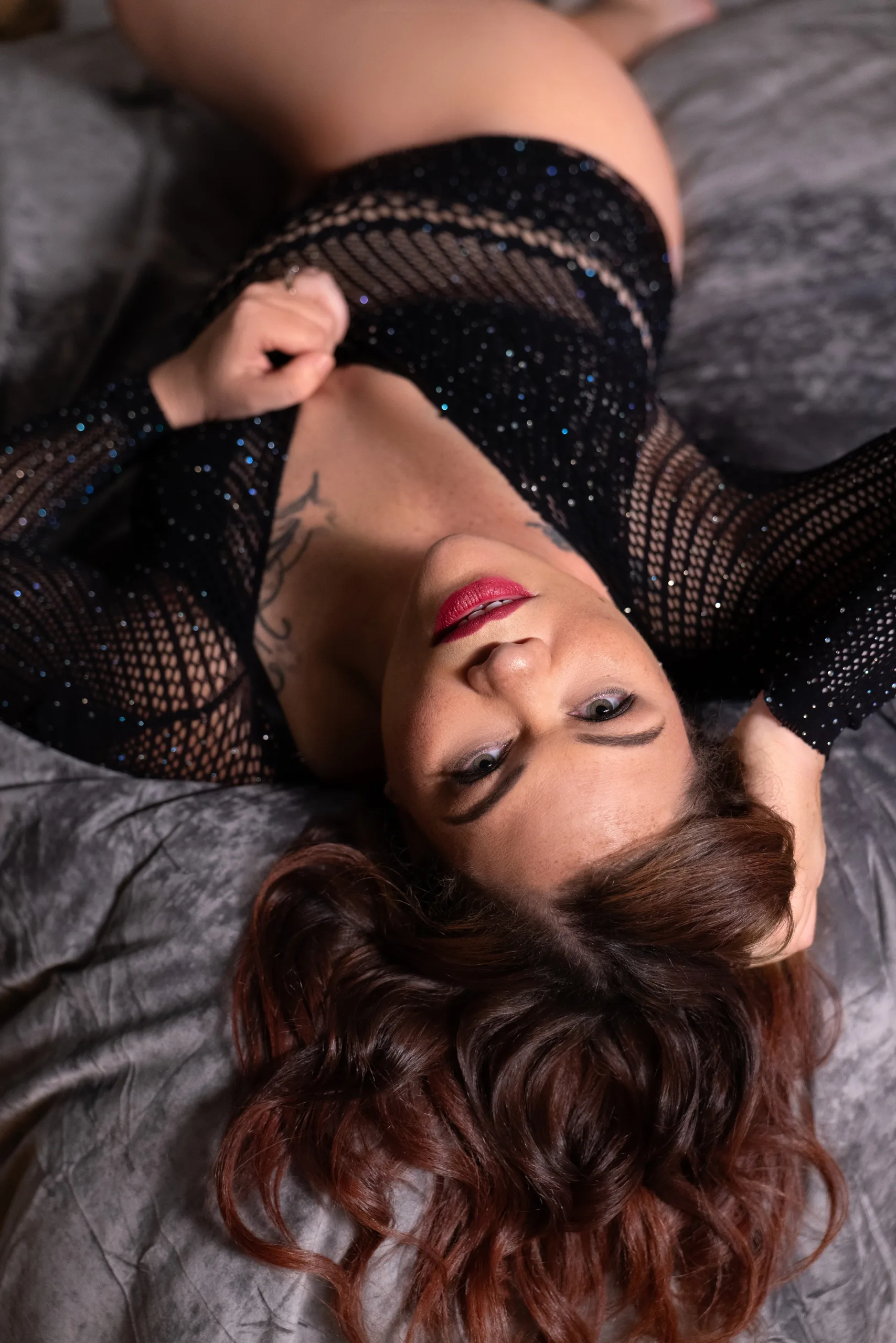 An adult in a black sparkly fishnet outfit lies on a gray surface with long wavy hair spread around her head.